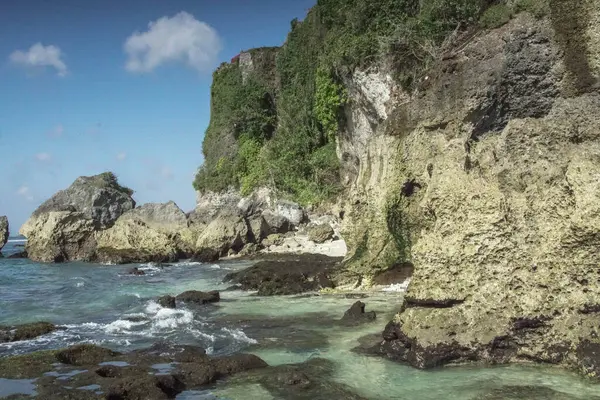 Yumuşak kum ve yosun kaplı kayaların üzerinde hafif dalgalarla yıkanan sakin bir tropikal plaj. Dramatik uçurumlar deniz manzarasını oluştururken, insanlar okyanusun tadını uzaktan çıkarırken görülebilir..