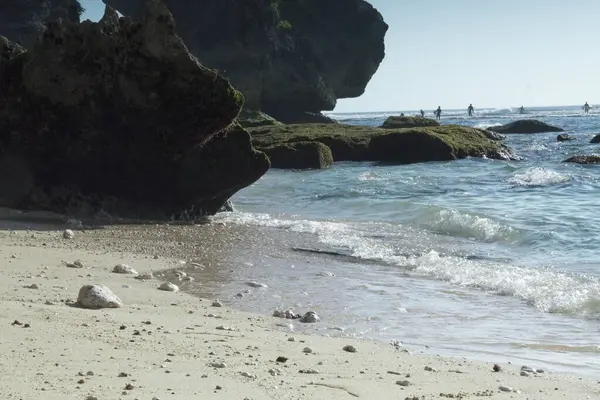 Yumuşak kum ve yosun kaplı kayaların üzerinde hafif dalgalarla yıkanan sakin bir tropikal plaj. Dramatik uçurumlar deniz manzarasını oluştururken, insanlar okyanusun tadını uzaktan çıkarırken görülebilir..