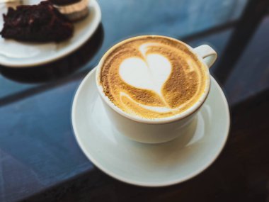Beyaz bardakta Latte sanat eseri ve arka planda çikolatalı kek. Kahve dükkanı atmosferi.