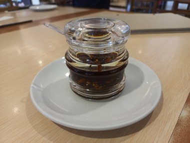 İçi canlı kırmızı biber yağıyla dolu berrak bir cam şişe. Eritilmiş biberlerin zengin rengini ve dokusunu sergiliyor. Şişe beyaz bir tabakta duruyor, basit ve iştah açıcı bir görüntü yaratıyor..