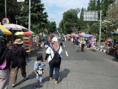 İnsanlar canlı bir Endonezya sokak pazarında şemsiyelerin altında renkli tezgahlarla dolaşıyorlar. Çocuklar da dahil olmak üzere bir aile, açık hava pazarının tadını çıkarıyor..