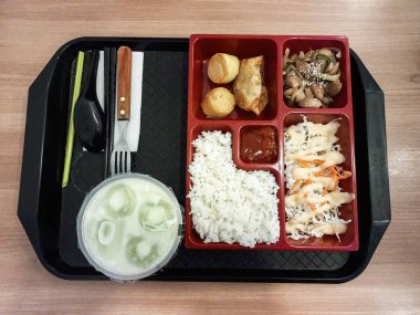 Bir tepside lezzetli bir teriyaki bento kutusu, içinde pilav, tavuk, gyoza ve salata, ferahlatıcı yeşil bir içecek ve kap kacak ile servis edilir..