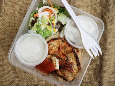 Plastik bir kabın içinde hazır yemek, ızgara tavuk bifteği, salata ve yanında patates püresi olan yakın çekim görüntüsü..