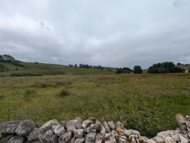 Geniş, sakin bir manzara bulutlu bir gökyüzünün altında sergileniyor, ön planda taştan bir duvar var, geniş bir alana açılıyor..