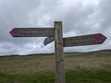 Ahşap bir işaret direği, bulutlu bir gökyüzünün altında Malham Cove ve Gordale 'e olan mesafeyi gösteren yürüyüş yollarını gösteriyor..