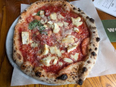 Taze malzemeli ahşap kaplı lezzetli bir pizza metal bir tabakta sunuluyor. Kabuğu çok güzel yanmış, malzemeler taze görünüyor..