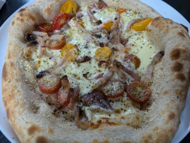 Beyaz bir tabakta taze kiraz domatesli, pastırmalı ve peynirli lezzetli bir pizza servis edilir. Köy pizzası mükemmel pişmiş, enfes bir yemek..