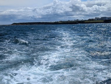 Uçsuz bucaksız mavi okyanus ufka doğru uzanıyor, bulutlu bir gökyüzünün altında uzak bir kıyı şeridi beliriyor, sakin bir deniz manzarası yaratıyor..
