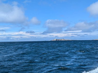 Çok güzel bir gökyüzünün altında engin bir okyanus. Uzakta deniz feneriyle kaplı küçük bir ada. Navigasyon ve tarihi işaret ediyor..