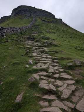 Bulutlu bir gökyüzünün altında zirveye doğru uzanan yemyeşil bir dağa doğru uzanan bir taş yol var. Bir taş duvar yürüyüşçülere rehberlik eder.
