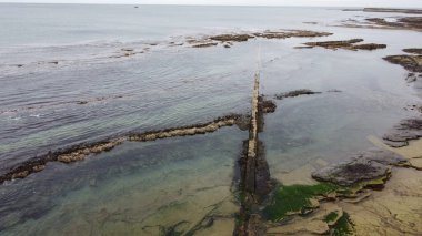 Sakin bir kıyı manzarası alçak bir gelgiti yakalar ve kıyı boyunca batık bir taş yapısını ve deniz yaşamını gözler önüne serer..
