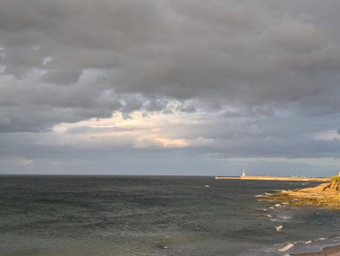 Karamsar bir gökyüzü altında, kara bulutlar ve uzak bir deniz feneri ile dramatik bir deniz manzarası yakalar. Dalgalar melankolik bir günde usulca sahile doğru yuvarlanıyor..