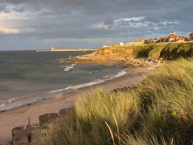 Sakin bir sahil manzarası, kumlu bir plaj, çimenli kum tepeleri ve kıyı şeridi boyunca antika evler. Deniz kasvetli bir gökyüzünün altında uzak bir iskeleye doğru uzanıyor..