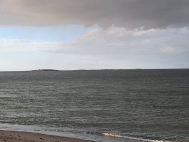 Dramatik bulutlar ve nazik dalgalarla dolu kasvetli bir gökyüzünün altında ufukta bir adanın olduğu sakin bir sahil manzarası..
