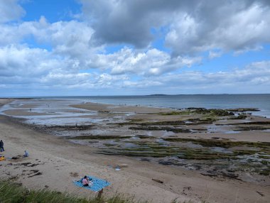 Resmedilmeye değer bir kıyı manzarası kumlu bir sahilin dramatik, bulutlu bir gökyüzünün altında insanların günün tadını çıkardığı bir sahili gösteriyor..