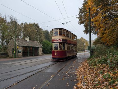 Kaliteli bir çift katlı tramvay, büyüleyici bir otobüs durağının yanındaki pitoresk bir sonbahar ortamında rayları boyunca yuvarlanıyor. Sahne sonbahar yapraklarıyla dolu..
