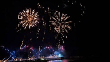 Havai fişekler gece gökyüzünü altın patlamaları ve renkli patikalarla aydınlatıyor. Suyun üzerindeki şenlikli bir kutlamada..