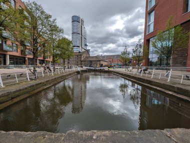 Şehir ortamında binaları ve bulutlu gökyüzünü yansıtan sakin bir kanal sahnesi. Su yüzeyi etrafındaki yapıları yansıtır..