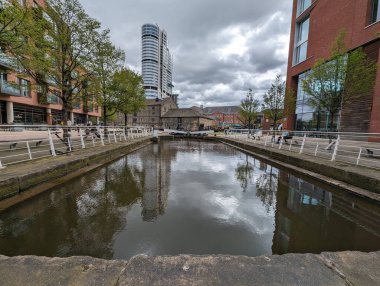 Modern binalarla çevrili bir kanal manzarası bulutlu gökyüzünü ve mimariyi yansıtıyor. İnsanlar yol boyunca yürür.