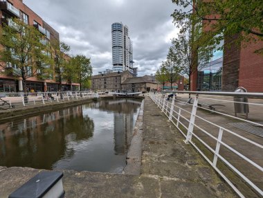 Bir kentsel kanal sahnesinde, suya yansıyan modern mimari yer alır. Bulutlu gökyüzü biraz atmosfer katıyor..