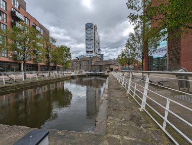 Şehirdeki sakin bir kanal manzarası binaları ve bulutlu bir gökyüzünü yansıtır. Modern mimari, su kenarındaki tarihi yapılarla çelişiyor..