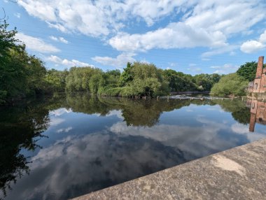 Sakin bir nehir manzarası kabarık beyaz bulutları ve kıyıları çevreleyen yemyeşil ağaçları yansıtır. Sakin bir yer..