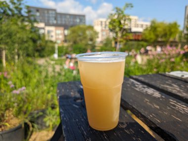 Serinletici bir bira şişesi dışarıdaki yıpranmış ahşap masada duruyor. İnsanlar bazı binaların yakınındaki güneşli günün tadını çıkarıyorlar..