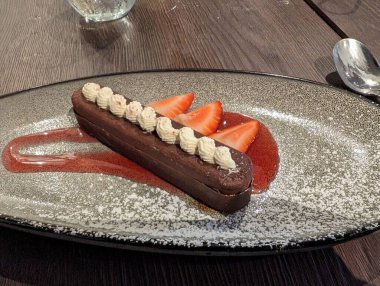 Süslü bir tabakta servis edilen krem şanti ve taze çilekle süslenmiş lezzetli bir çikolatalı tatlı. Sunumda canlı bir çilek sosu ve toz haline getirilmiş şeker yer alıyor..