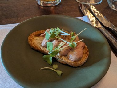 Bir gurme aperatifi, bir restoranda koyu yeşil bir tabağın üzerinde kızarmış baget diliminde mikro yeşillikle ezme sunar..