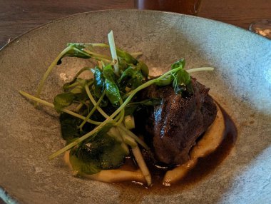 Canlı bir bezelye filizi garnitürü ile tamamlanmış yumuşak bir et parçasından oluşan bir gurme tabağı pürüzsüz bir püre yatağında servis edilir..