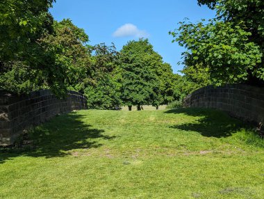 Canlı bir manzara, yemyeşil çimenler, taş duvarlar ve mavi gökyüzünün altındaki ağaçlar sükuneti çağrıştırıyor..