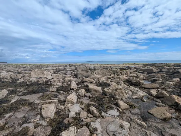 Manzaralı bir kıyı manzarası, bulutlu bir gökyüzünün altında ufka doğru uzanan kayalık bir genişliği ve görülebilir adaları gösteriyor..