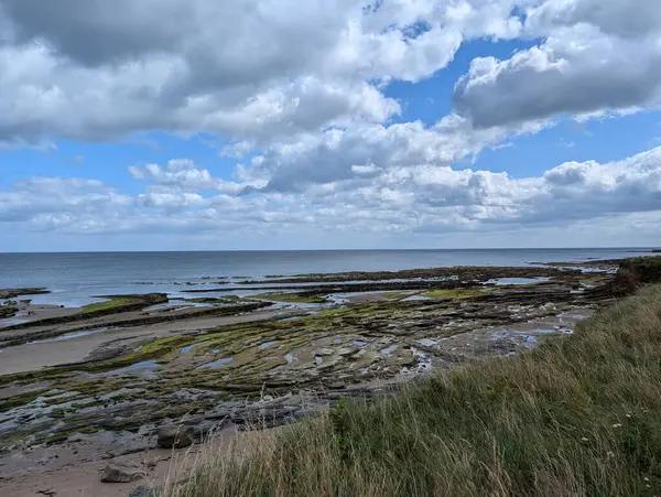 Okyanus manzarası, kumlu plaj, kaya oluşumları ve geniş bulutlu gökyüzü, dingin ve sakinleştirici..