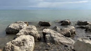 Yumuşak göl dalgaları, sakin bir kıyı şeridinde kayalara çarpıyor. Uzaktan dağ manzaralı. Huzurlu, doğal bir atmosfer yaratıyor.