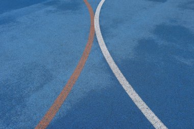 Canlı mavi sahada kavisli turuncu ve beyaz spor çizgileri, yağmurdan sonra dokulu ıslak kauçuk yüzey üzerinde soyut basketbol veya atletik işaretler