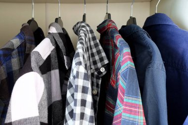 Bir gardıropta plastik askılarda asılı çeşitli ekose ve katı gömlekler. Dolapta düzenlenmiş renkli flanel ve pamuklu giysiler. Moda, kostüm organizasyonu ve erkek giyim konsepti