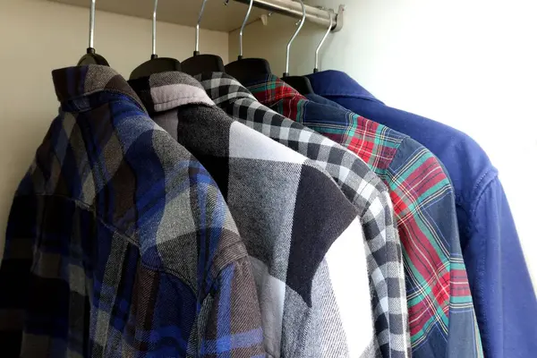 Ahşap bir gardıropta siyah askılara asılı sıradan ve koyu renkli gömlekler. Parlak gün ışığı, flanel desenli ve renk kontrastlı düzenli erkek kıyafetleri.