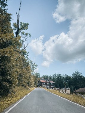 Sakin, dar bir yol, ağaçların arasına kurulmuş büyüleyici bir kır evine giden ormanın içinden yavaşça yukarı doğru esiyor. Parçalı bulutlu gökyüzü ve doğal çevre huzurlu bir kırsal atmosfer yaratıyor..