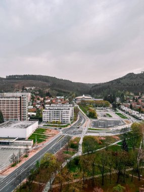 Yeşil tepeler ve sık ağaçlıklar tarafından çerçevelenmiş sakin bir şehir bölgesinin havadan görünüşü. Kentsel alanda modern konut binaları, geniş yollar ve bulutlu bir gökyüzü altında huzurlu bir yerleşim yeri yer alıyor..