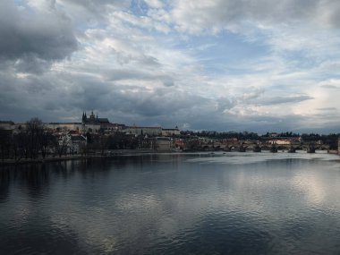 Prag Kalesi ve Vltava Nehri 'ni kaplayan Charles Köprüsü' nün karamsar bir gökyüzünün altında suya yansıyan görüntüsü. Prag 'ın tarihi ufuk çizgisi, başımızın üstündeki dinamik bulutlarla çelişiyor ve zamansız bir güzellik hissi uyandırıyor..