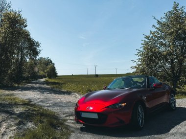 Parlak mavi gökyüzünün altında yeşil tarlalar ve ağaçlarla çevrili, üstü açık, parlak kırmızı Mazda MX5 model bir araba. Bir yaz gezisi ya da araba meraklıları için mükemmel bir sahne..