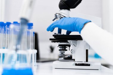 Mikroskop kullanarak bir araştırmacıyı ya da tıp uzmanını gösteren, hassas ve bilimsel araştırmayı vurgulayan bir laboratuvar sahnesinin yakın çekimi. Mavi eldivenli eller test sırasında mikroskobu ayarlar