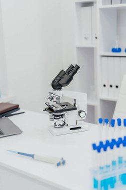 Temiz beyaz bir masa üzerinde yüksek güçlü bir mikroskop bulunan modern bir laboratuvar düzeneği. Mikroskobun etrafında mavi sıvıyla dolu test tüpleri ve aktif olduğunu gösteren bir pipet var.