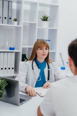 Bir kadın doktor tıbbi danışma sırasında bir hastayı dikkatle dinler. Laboratuvar ekipmanları ve organize raflarla kurulmuş ofis ayarları profesyonel bir sağlık ortamını yansıtıyor