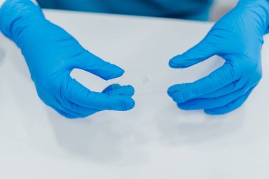 Eldivenli ellerin dikkatle tuttuğu bir mikroskop kaydırağını bir damla sıvıyla dikkatle tutması laboratuvar çalışmalarında hassasiyet ve temizliği vurguluyor. Parlak mavi eldivenler beyaz eldivenlerle tezat oluşturuyor.