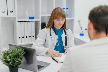Profesyonel bir kadın doktor muayene sırasında hasta kayıtlarını dikkatle inceler. Bir dizüstü bilgisayar, laboratuvar aletleri ve organize raflar ile donatılmış sağlık ofisi, modern ve