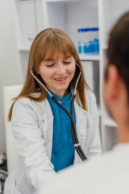 Steteskop takan gülümseyen bir kadın doktor muayene sırasında bir hastayı dinler. Sahne, klinik ortamda profesyonel ve şefkatli bir sağlık hizmeti etkileşimi yakalıyor. Yüksek kalite