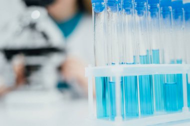 Laboratuvar deneylerinin hassasiyetini ve odak noktasını vurgulayan mavi sıvı içeren test tüpleriyle dolu bir rafın yakın plan çekimi. Bulanık arka plan bir bilim adamının...