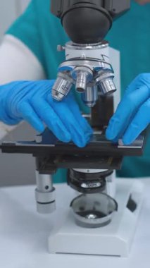 Mavi üniformalı ve eldivenli odaklanmış bir laboratuvar teknisyeni modern bir laboratuvarda bilimsel örnekler hazırlıyor. Görüntü ön planda bir mikroskop, test tüpleri ve temiz, düzenli