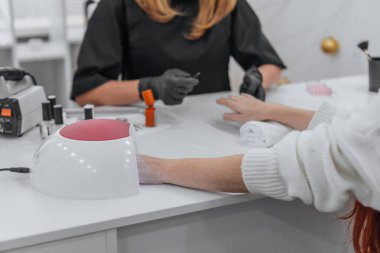 Profesyonel bir kuaför salonunda jöle cilası için ultraviyole lambanın altına el süren bir müşteri. Siyah eldiven giyen manikürcü arka planda çalışıyor, temiz ve modern bir çalışma alanı sergiliyor